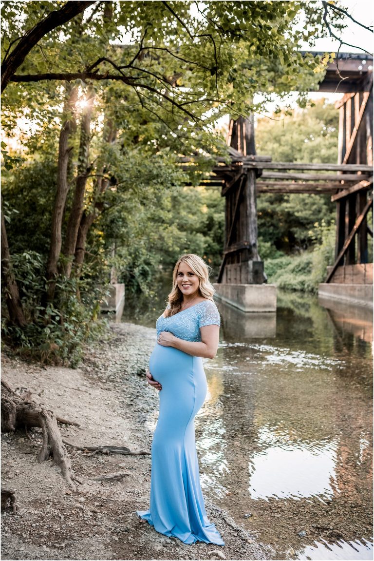 Stunning expecting mother standing near creek in Cedar Park Texas wearing a blue maxi dress during maternity photoshoot by natural light portrait photographer